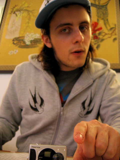 A young man, wearing a cap and hoodie-style jacket, looks askance at the camera to which he points.